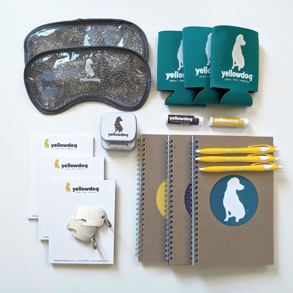 An assortment of promotional items: sleep masks with microbead filling, koozies, mints, notepads, chap sticks, journals, and pens all customized with YellowDog branding.