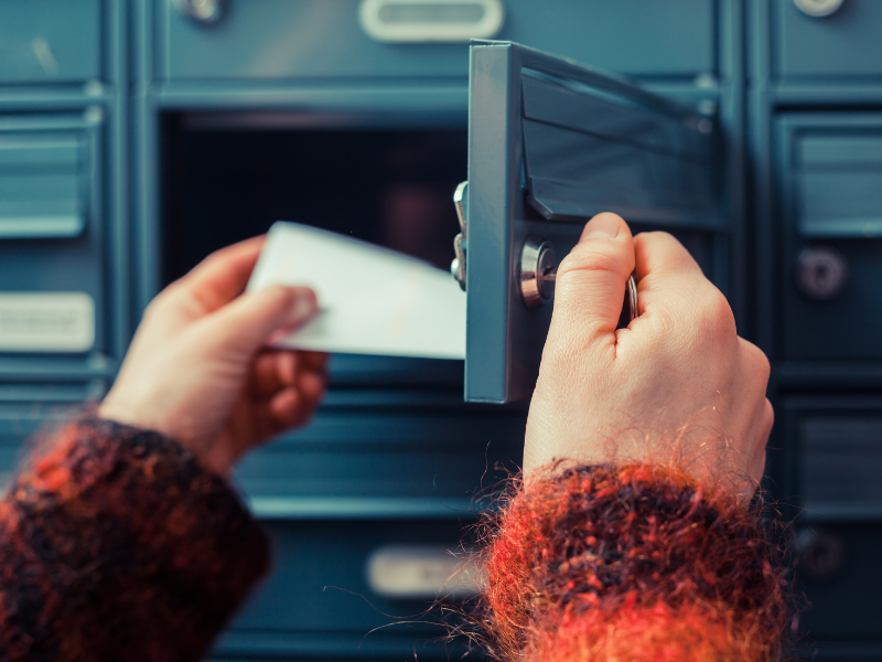 Two hands with fuzzy orange sleeves are shown unlocking a mailbox in a wall of mailboxes, pulling out a piece of direct mail. 