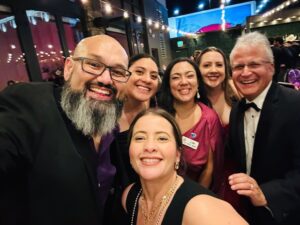 Group selfie of six adults in formal attire, smiling outside at night under string lights.