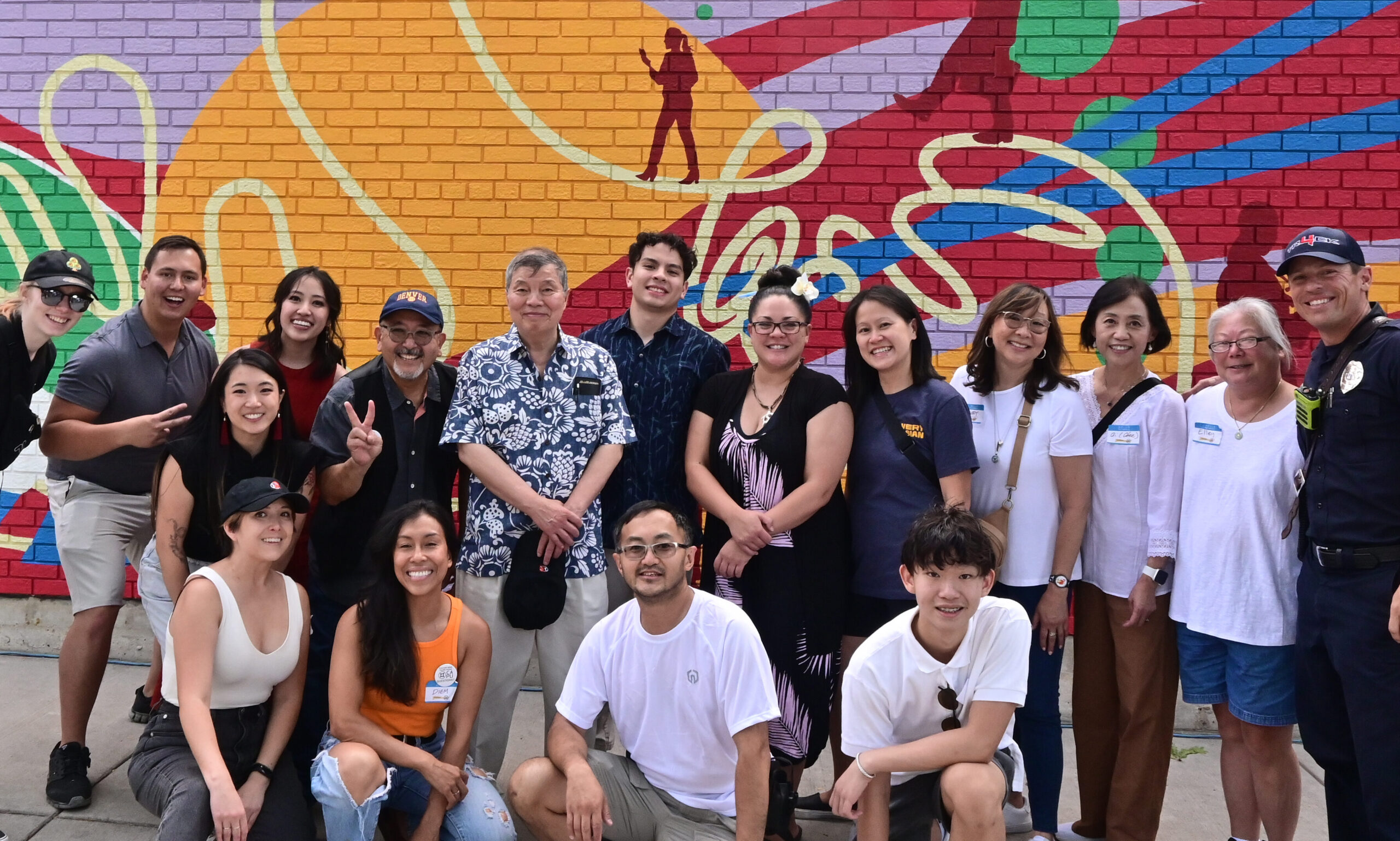 A group of 17 adults smiling at the camera in front of a brightly painted brick wall.