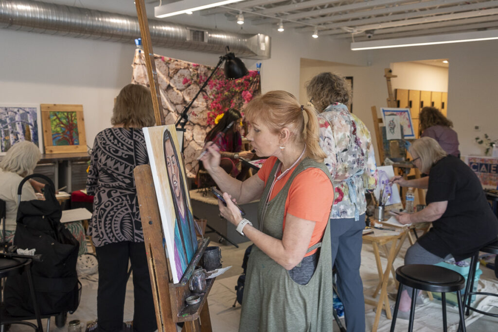 A woman in an orange t-shirt and green cover-alls painting a canvas.