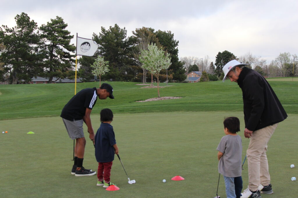 Two adults showing two small children how to putt on a golf course.