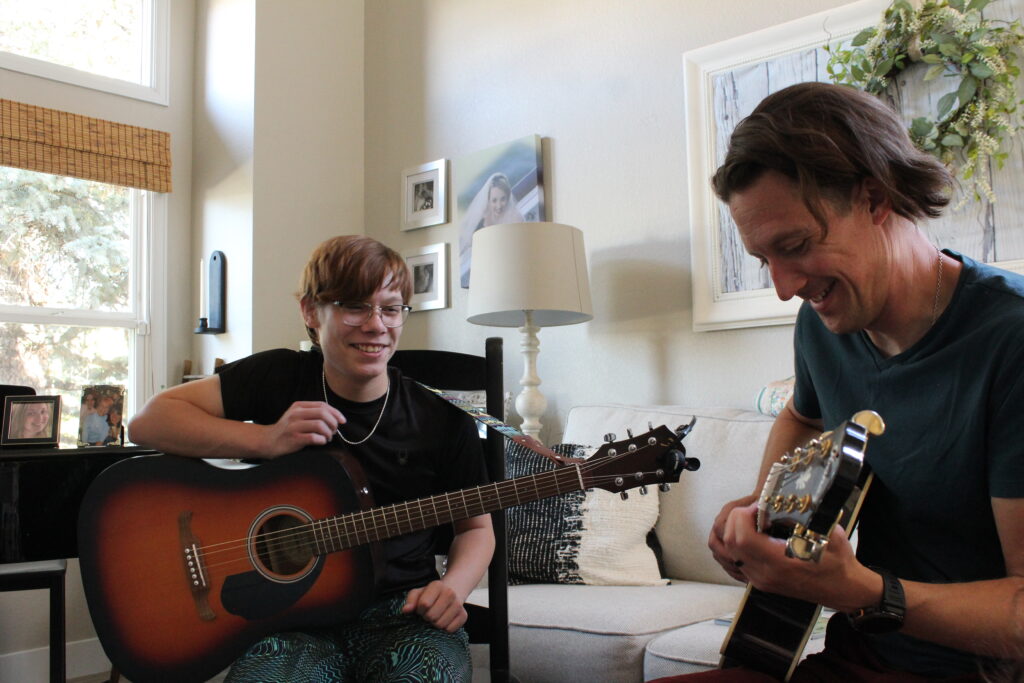 A young man and an adult man playing guitars in a white living room.