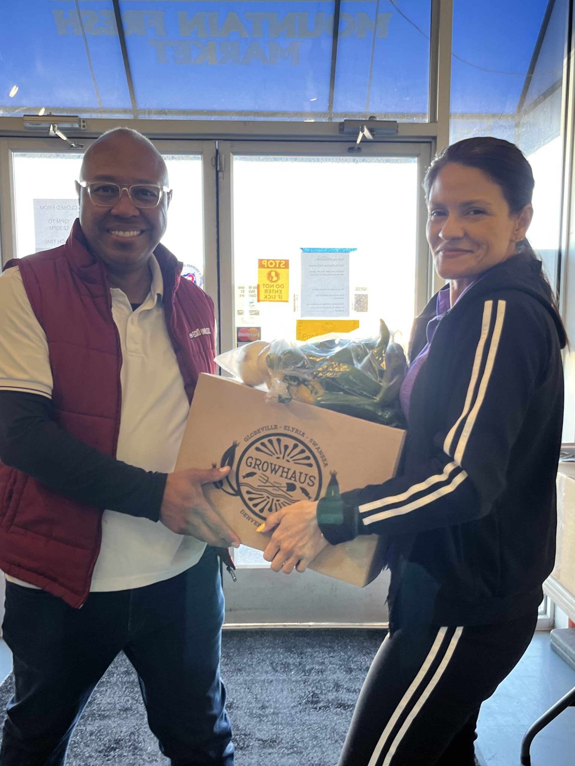 Two adults holding a cardboard box full of vegetables with the GrowHaus logo on the side, both smiling at the camera.