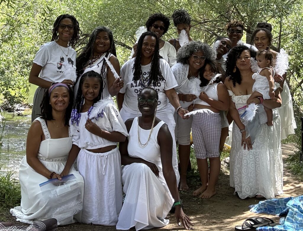 A group of women/femmes, all dressed in white, posing next to a river, surrounded by green trees.