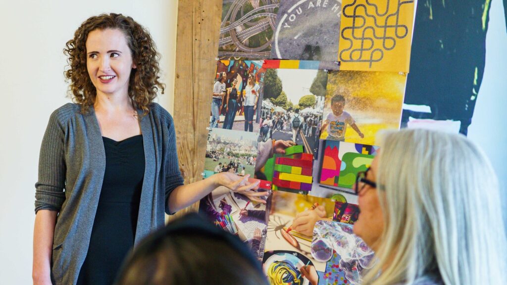 A mood board can help communicate your vision as part of your creative brief. Here, a young woman with curly hair presents a community event mood board to a team of designers.
