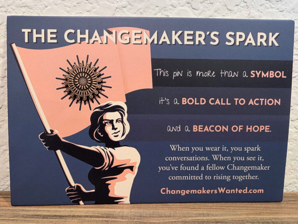 A spark-shaped lapel pin on a navy, cream and white card, depicting a graphic of a woman waving a flag, and the words: "The Changemaker's Spark - This pin is more than a symbol. It's a bold call to action, and a beacon of hope. When you wear it, you spark conversations. When you see it, you've found a fellow Changemaker commited to rising together. ChangemakersWanted.com"