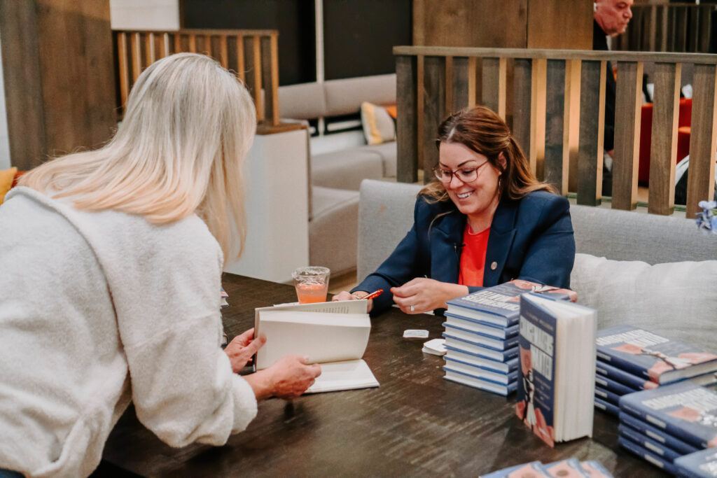 A woman wearing white leaning over a table over an open book, toward a woman wearing a blue suit, seated behind the table, signing books.