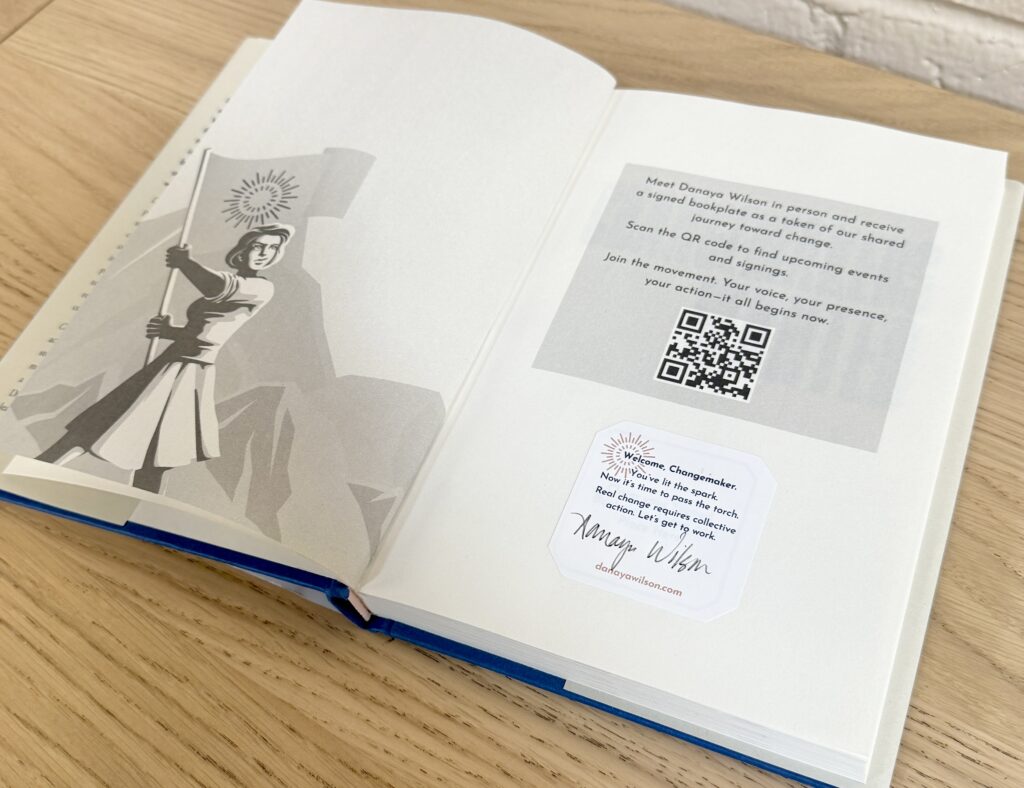 An open book displays a graphic illustration of a woman waving a flag in front of mountains, with a personalized name plate on the right side.