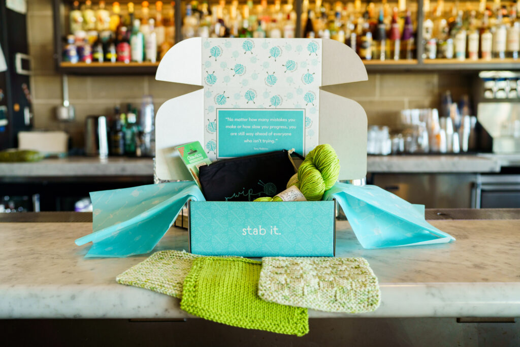 Open WIP kit: desinged teal box with the words "stab it" on the front panel, sitting open on a table with branded tissue paper, yarn, tools and canvas zip bag on display.